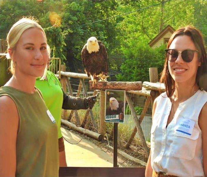 two women smile at the camera with an eagle on a perch in the background