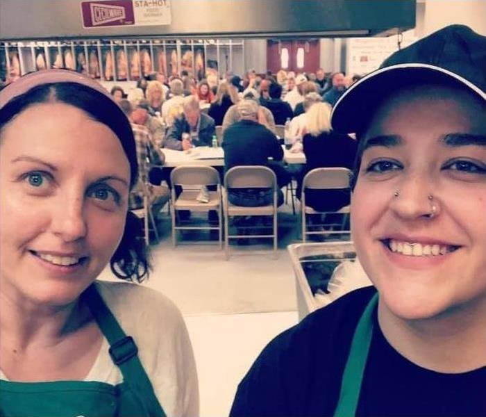 two women with hats and aprons in front, in the background there are people sitting in chairs in a fire hall 