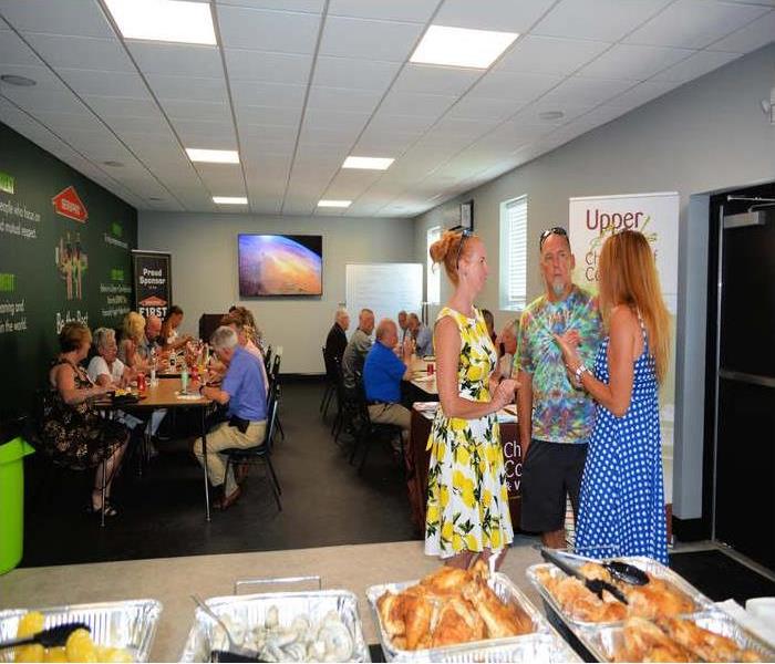 A food table in the forefront, three people standing up talking towards the front and others sitting at tables in the back