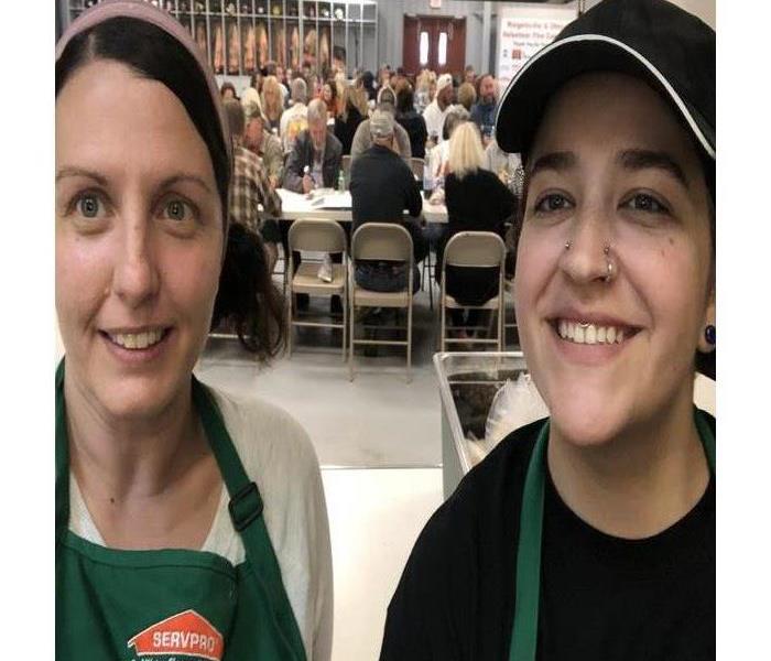 Two women standing inside a kitchen with a serving window behind them people sit at tables playing bingo.